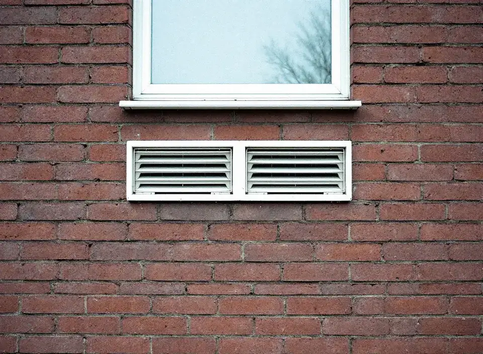 Twee witte roosters naast elkaar in een bakstenen gevel, vlak onder een raam: de openingen van een airco zonder buitenunit